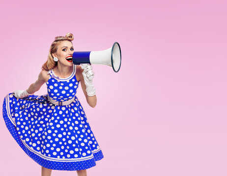 Image of woman holding megaphone, dressed in pin up style blue dress in polka dot and white gloves, on pink background. Caucasian blond model posing in retro fashion vintage studio concept.