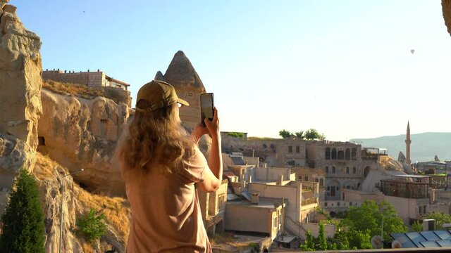 Woman stands on balcony or terrace of cave hotel and takes mobile pictures and videos of amazing unusual landscape of Cappadocia, Turkey using cell phone. Hot air balloons flying in blue sky