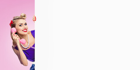 Image of woman with phone, in pin up style dress, showing blank signboard with copyspace area, over pink background. Caucasian blond model posing in retro fashion and vintage studio concept.