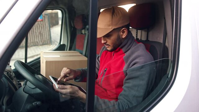 Delivery boy wearing a cap sitting in the Driving seat of the van with a cardboard box placed on the other seat. Looking up address using a tablet. Concept of parcel delivery services. 