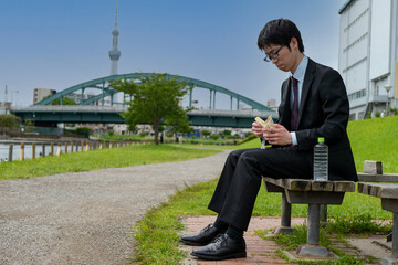 東京スカイツリーをバックに公園でサンドイッチを食べるビジネスマン