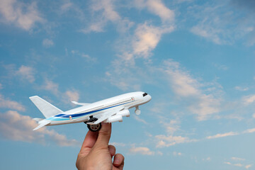 A hand holding a toy white airplane against the backdrop of a beautiful sky with clouds, copy space. Travel, vacation concept. Post-pandemic flight concept