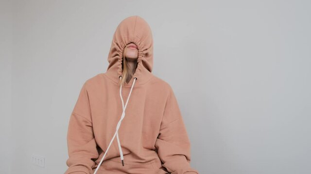 Sad teenage girl with large birthmark on face hides in hood lacing up beige sweater sitting on light grey background