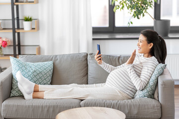 pregnancy, rest, people and expectation concept - happy smiling pregnant asian woman sitting on sofa at home having video call on smartphone