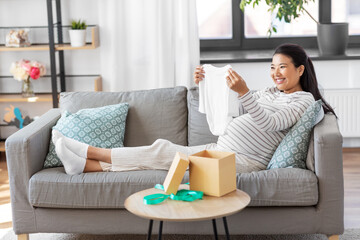 pregnancy, holidays and people concept - happy smiling pregnant asian woman opening gift box with baby's bodysuit at home
