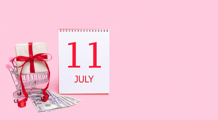 A gift box in a shopping trolley, dollars and a calendar with the date of 11 july on a pink background.