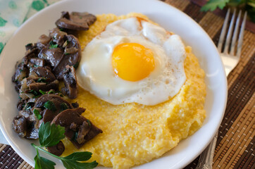 Corn porridge, fried egg, fried mushrooms with onions in a plate on a wicker bamboo napkin.