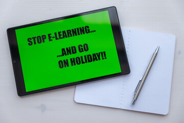 Tablet showing black text on green background "Stop e-learning, and go on holiday", next to white notebook, pen, on white wooden surface. 
