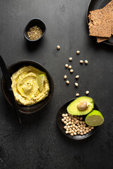 Black ceramic bowl with hummus on black backround with chickpea beans, spices, avocado, lime and multigrain bread,