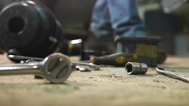 Car auto repair shop. Repairing vehicle garage your own home. Man takes screwdriver, tightens screw, gloved hand picks up the wrenches from concrete floor. Active work in full swing, mess tools floor