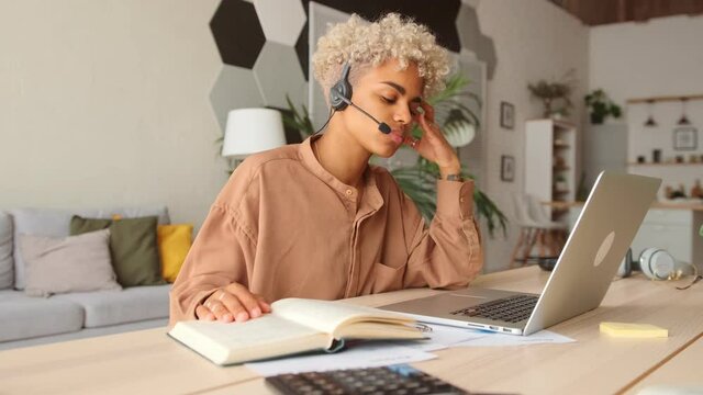 Young dark skinned female has blonde curly hair wear headset talk by conference call speak looking at laptop at home office video chat job interview or distance language course class as online teacher