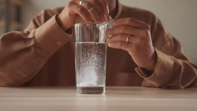 Sick ill Afro woman holding dissolving effervescent aspirin pill drop tablet into glass of sparkling water taking painkiller medicine to relieve headache pain healthcare medicament close up view
