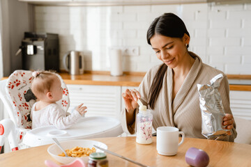 Young brunette mother smiling and feeding her baby in kitchen at home