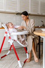 Fototapeta premium Young brunette mother smiling and feeding her baby in kitchen at home