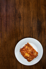 Lasagna in Bolognese sauce, made with ham cheese, tomato sauce and ground beef. Served on a White Plate. Top view photo. Dark background wood.