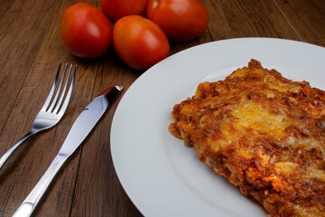 Lasagna in Bolognese sauce, made with ham cheese, tomato sauce and ground beef. Lasagna served on the plate. Fork and knife on the table, raw tomatoes in the background.