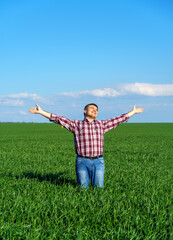 Naklejka na ściany i meble a man as a farmer poses in a field, dressed in a plaid shirt and jeans, he looks into the distance and raises his hands high in the sun