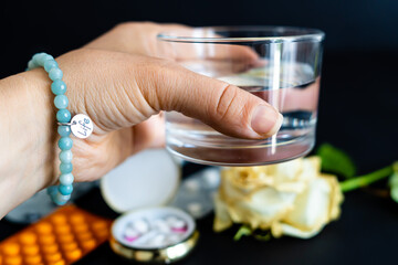Mano che tiene un bicchiere d'acqua in primo piano e un porta pillole e altre medicine sfuocate sullo sfondo e una rosa bianca. Prendere le medicine. Salute.