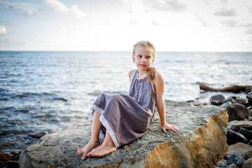 A blonde girl in a sundress sits on a stone on the seashore.