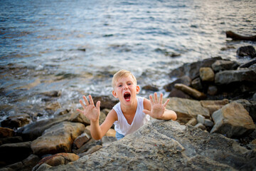 A boy in a white T-shirt and denim shorts peers out from behind a large stone at the seashore.