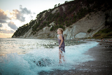 Fototapeta premium A girl in a purple sundress stands on the surf line against the background of the sea and rocks