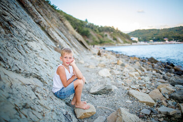 A boy in a white T-shirt and denim shorts sits on a rock near the seashore
