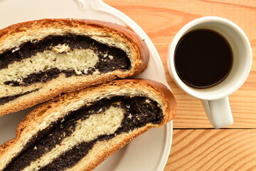 Two slices of freshly baked poppy seed roll, on a white dish with a cup of coffee on a wooden table, close-up, top view.