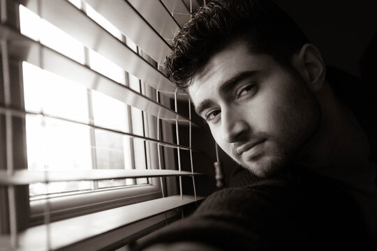Portrait of handsome man with beard sitting next to window with blinds, looking at camera
