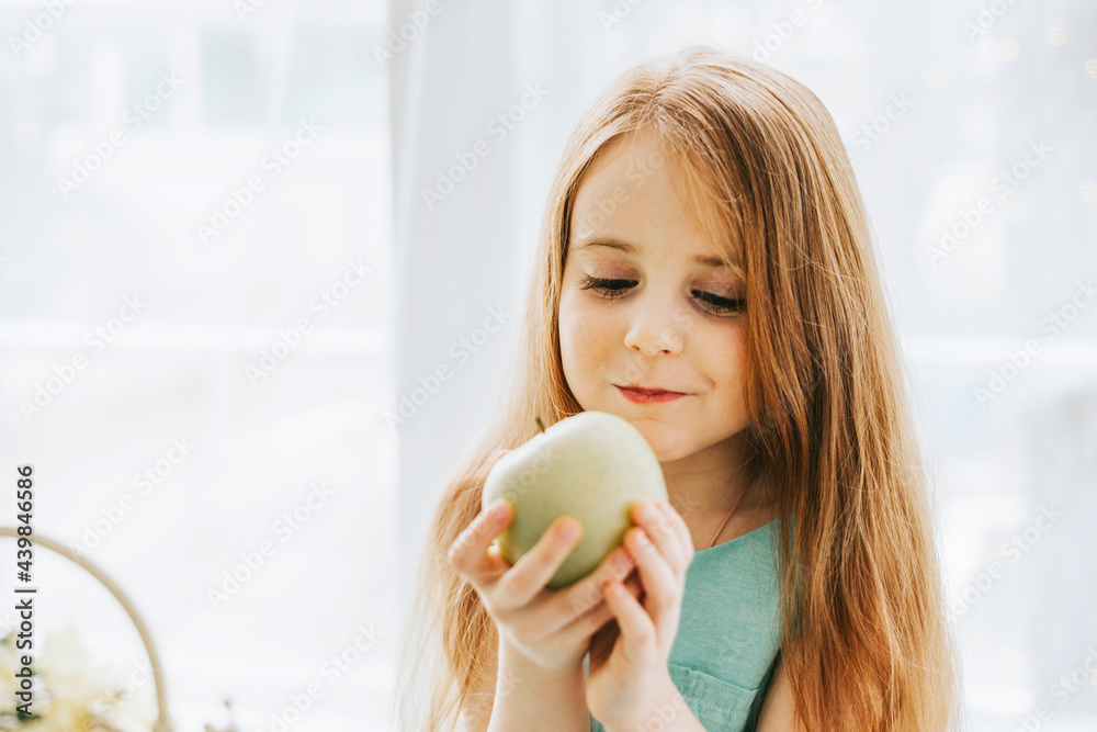 Wall mural little red haired girl is sitting in the kitchen of a country house eating an apple next to the sink where the ducklings are bathing