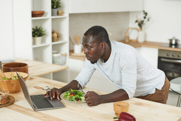 Fototapeta na wymiar Young man eating vegetable salad and using laptop in the kitchen