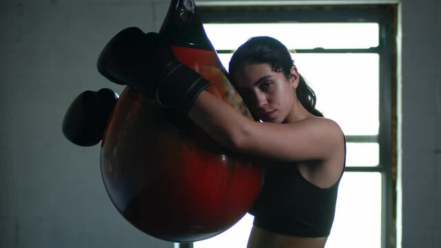 Tired and sweaty woman fighter leaning on the punching bag after intensive trining and looking in the camera.
