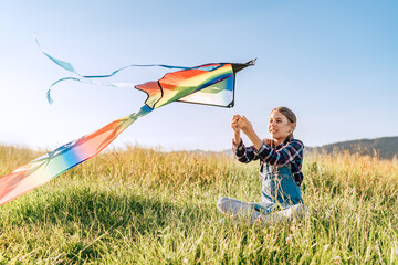 9YO smiling girl sitting on the grass and preparing colorful rainbow kite toy for flying. Happy...