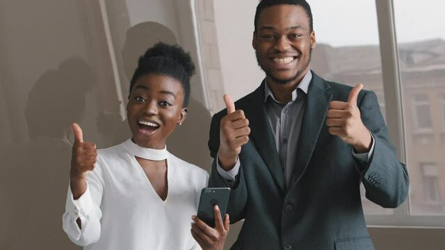 Emotional happy cheerful young African ethnic colleagues looking at telephone screen, laughing feeling excited of getting message with amazing news, celebrating online lottery win showing thumbs up.