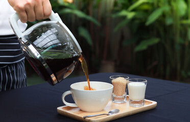 Pour coffee into a cup on a wooden tray. placed on a table covered with a black tablecloth. The atmosphere in the garden of the restaurant with blurred green leaves background.
