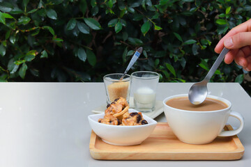 Part of hand holding spoon to stir coffee and there are cookies, sugar and milk together on wooden tray. Garden atmosphere.
