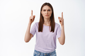 People emotions. Young woman pointing up and frowning displeased, dislike banner, condemn announcement, disapppointed by something bad, white background