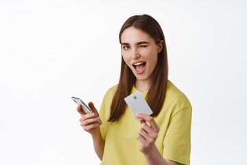Cheerful girl pays on phone, shopping online with smartphone, showing credit card and winking, got...