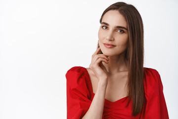 Close up of natural, alluring woman with sensual smile, romantic make up and red passionate dress, gazing at camera, touching facial skin, looking thoughtful, white background