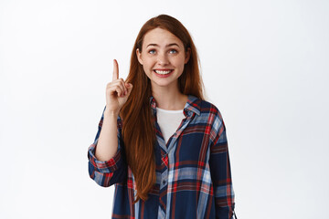 Cheerful teen redhead girl, smiling and ponting looking up, staring at top logo with amazed happy face, standing against white background