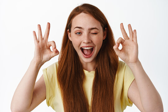 Close Up Portrait Of Redhead Positive Girl, Winking And Showing Okay OK Sign, No Problem, Praise Good Thing, Well Done, Great Job, Standing Over White Background