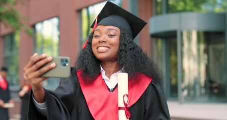 Portrait of a happy attractive multiracial graduate holding her smartphone and chatting via video call. College university graduation day. Proud woman towards camera with big smile
