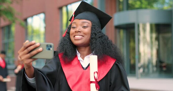 Portrait of a happy attractive multiracial graduate holding her smartphone and chatting via video call. College university graduation day. Proud woman towards camera with big smile