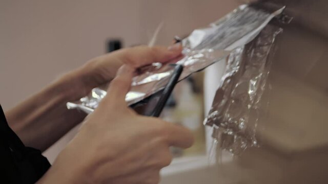 Close-up of the process of highlighting the long brown hair of a girl. The hairdresser prepares the strong beautiful hair of the girl for highlighting, combing the hair, wrapping wet hair in foil