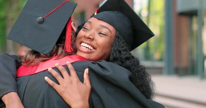 Pretty diverse graduates are embracing with diploma and rejoicing with each other. Young woman is hugging her groupmate with other students moving and talking in background