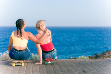 Girls sit on their skateboards near of the beach. Young sporty friends having fun in summer vacation. Travel, holidays and healthy lifestyle concept.