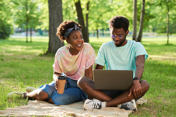 Naklejka na ściany i meble African young couple sitting on the grass drinking coffee and using laptop computer in the park