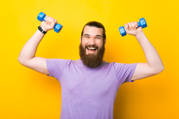 Portrait of happy cheerful bearded man working with small dumbbells over yellow background