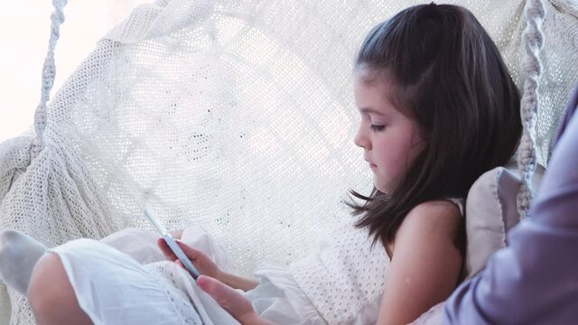 Curious cute preschool girl using digital tablet technology device sitting in comfortable circle chair alone. Small child hold pad computer surfing internet play game at home. Tech addiction concept