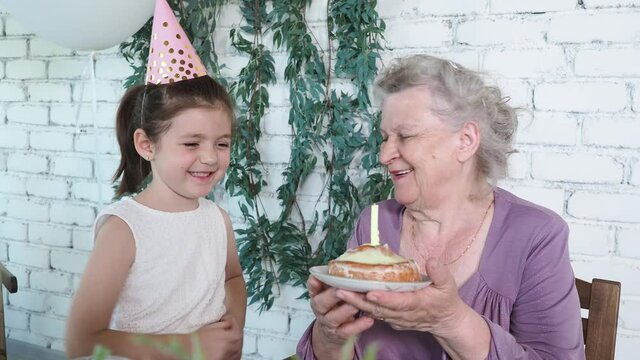 Happy smiling little cute girl and senior grandmother celebrate 85th birthday, holds cake and blowing out candle together, white brick background. Enjoy, respect and warm relation. Celebration concept