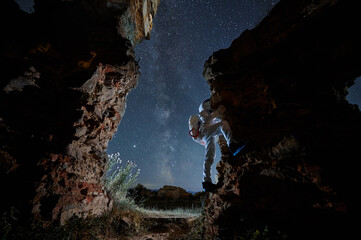 Spaceman in white suit and helmet is climbing on rock on background of starry sky at night. Man in...